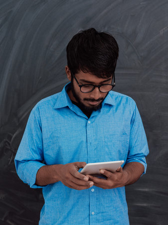 Indian smiling young student in blue shirt and glasses using tablet, smartphone and posing on school...