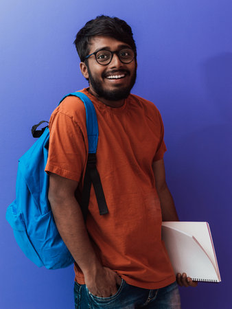 Indian student with blue backpack, glasses and notebook posing on purple background. The concept of...