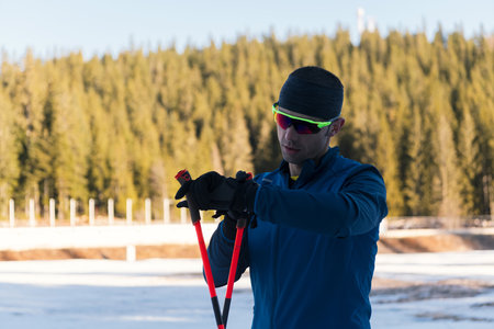 Handsome male athlete with cross country skis, taking fresh breath and having break after hard workout training in a snowy forest. Checking smartwatch. Healthy winter lifestyle.の写真素材