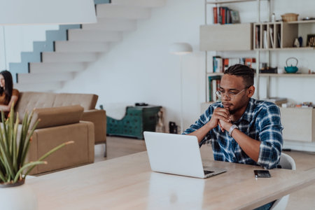 African American man in glasses sitting at a table in a modern living room, using a laptop for business video chat, conversation with friends and entertainmentの写真素材