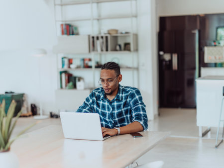 African American man in glasses sitting at a table in a modern living room, using a laptop for business video chat, conversation with friends and entertainmentの写真素材