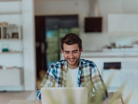 The man sitting at a table in a modern living room, with headphones using a laptop for business video chat, conversation with friends and entertainmentの写真素材