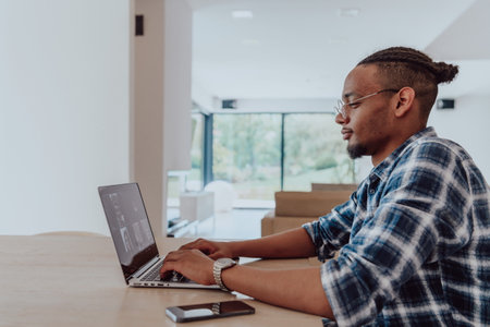 African American man in glasses sitting at a table in a modern living room, using a laptop for business video chat, conversation with friends and entertainmentの写真素材