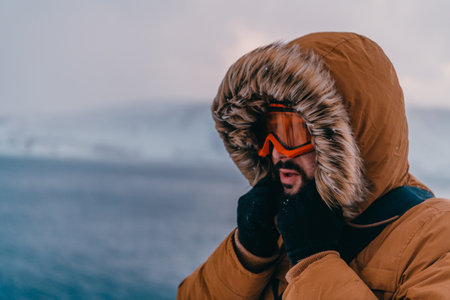 Headshot photo of a man in a cold snowy area wearing a thick brown winter jacket, snow goggles and gloves. Life in cold regions of the country.の写真素材