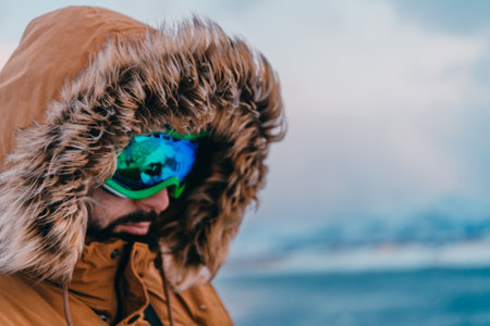 Headshot photo of a man in a cold snowy area wearing a thick brown winter jacket, snow goggles and gloves. Life in cold regions of the country.の写真素材