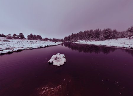 An aerial view of a frozen river flowing through snow-covered forests on a cloudy sunset sky...