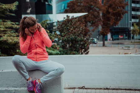 a woman in a sports outfit is resting in a city environment after a hard morning workout while using noiseless headphonesの写真素材