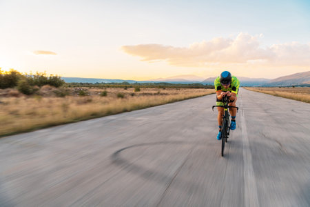 Triathlete riding his bicycle during sunset, preparing for a marathon. The warm colors of the sky provide a beautiful backdrop for his determined and focused effort.の写真素材