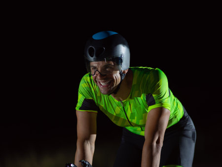 A triathlete rides his bike in the darkness of night, pushing himself to prepare for a marathon. The contrast between the darkness and the light of his bike creates a sense of drama and highlights the athletes determination and perseverance.の写真素材