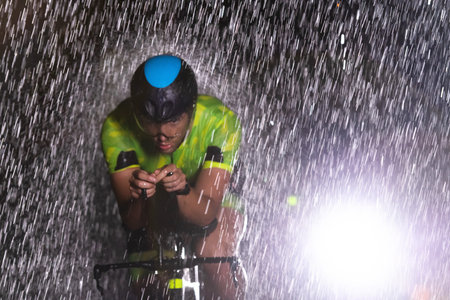 A triathlete braving the rain as he cycles through the night, preparing himself for the upcoming marathon. The blurred raindrops in the foreground and the dark, moody atmosphere in the background add to the sense of determination and grit shown by the athlete.の写真素材