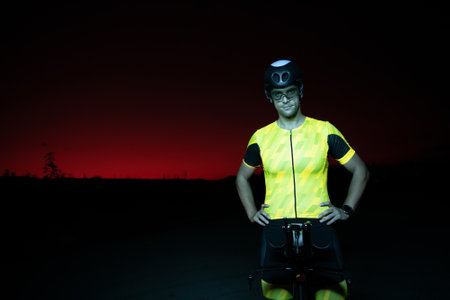 A triathlete resting on the road after a tough bike ride in the dark night, leaning on his bike in complete exhaustionの写真素材
