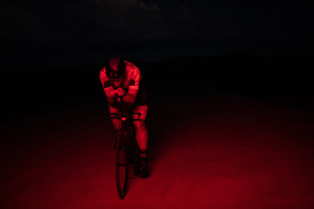 A triathlete rides his bike in the darkness of night, pushing himself to prepare for a marathon. The contrast between the darkness and the light of his bike creates a sense of drama and highlights the athletes determination and perseverance.の写真素材