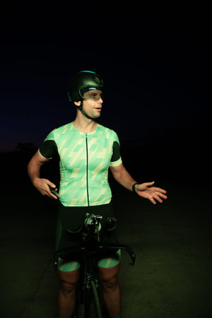 A triathlete resting on the road after a tough bike ride in the dark night, leaning on his bike in...
