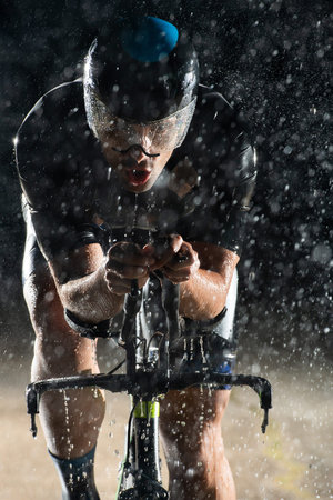 A triathlete braving the rain as he cycles through the night, preparing himself for the upcoming marathon. The blurred raindrops in the foreground and the dark, moody atmosphere in the background add to the sense of determination and grit shown by the athlete.の写真素材