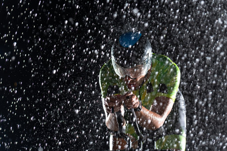 A triathlete braving the rain as he cycles through the night, preparing himself for the upcoming marathon. The blurred raindrops in the foreground and the dark, moody atmosphere in the background add to the sense of determination and grit shown by the athlete.の写真素材