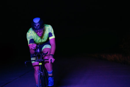 A triathlete rides his bike in the darkness of night, pushing himself to prepare for a marathon. The contrast between the darkness and the light of his bike creates a sense of drama and highlights the athletes determination and perseverance.の写真素材