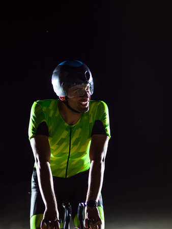 A triathlete rides his bike in the darkness of night, pushing himself to prepare for a marathon. The contrast between the darkness and the light of his bike creates a sense of drama and highlights the athletes determination and perseverance.の写真素材