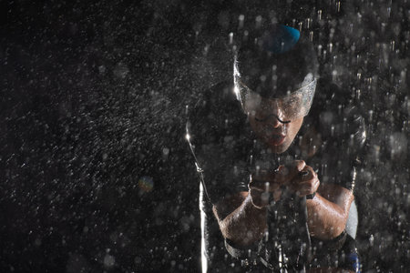 A triathlete braving the rain as he cycles through the night, preparing himself for the upcoming marathon. The blurred raindrops in the foreground and the dark, moody atmosphere in the background add to the sense of determination and grit shown by the athlete.の写真素材