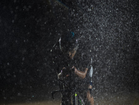 A triathlete braving the rain as he cycles through the night, preparing himself for the upcoming marathon. The blurred raindrops in the foreground and the dark, moody atmosphere in the background add to the sense of determination and grit shown by the athlete.の写真素材