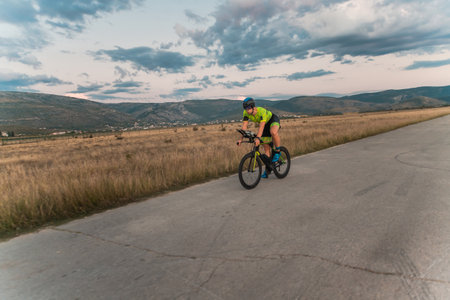 Triathlete riding his bicycle during sunset, preparing for a marathon. The warm colors of the sky provide a beautiful backdrop for his determined and focused effort.の写真素材