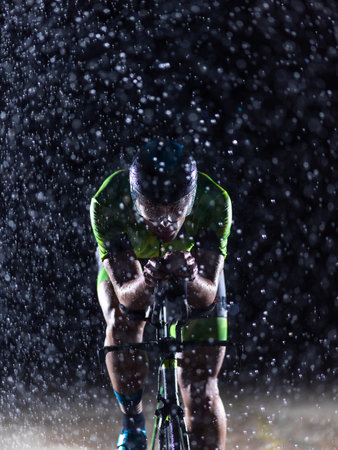 A triathlete braving the rain as he cycles through the night, preparing himself for the upcoming marathon. The blurred raindrops in the foreground and the dark, moody atmosphere in the background add to the sense of determination and grit shown by the athlete.の写真素材