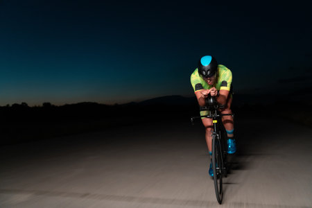 A triathlete rides his bike in the darkness of night, pushing himself to prepare for a marathon. The contrast between the darkness and the light of his bike creates a sense of drama and highlights the athletes determination and perseverance.の写真素材
