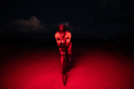 A triathlete rides his bike in the darkness of night, pushing himself to prepare for a marathon. The contrast between the darkness and the light of his bike creates a sense of drama and highlights the athletes determination and perseverance.の写真素材