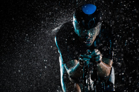 A triathlete braving the rain as he cycles through the night, preparing himself for the upcoming marathon. The blurred raindrops in the foreground and the dark, moody atmosphere in the background add to the sense of determination and grit shown by the athlete.の写真素材