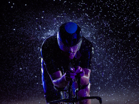 A triathlete braving the rain as he cycles through the night, preparing himself for the upcoming marathon. The blurred raindrops in the foreground and the dark, moody atmosphere in the background add to the sense of determination and grit shown by the athlete.の写真素材