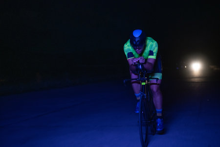 A triathlete rides his bike in the darkness of night, pushing himself to prepare for a marathon. The contrast between the darkness and the light of his bike creates a sense of drama and highlights the athletes determination and perseverance.の写真素材