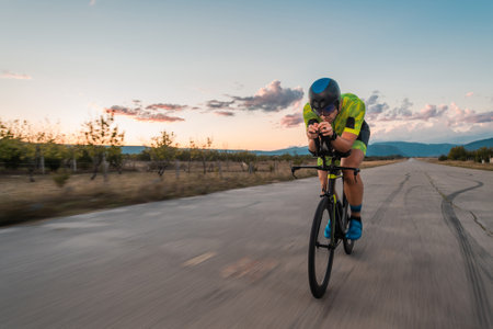 Triathlete riding his bicycle during sunset, preparing for a marathon. The warm colors of the sky provide a beautiful backdrop for his determined and focused effort.の写真素材