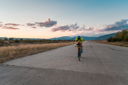 Triathlete riding his bicycle during sunset, preparing for a marathon. The warm colors of the sky provide a beautiful backdrop for his determined and focused effort.の写真素材