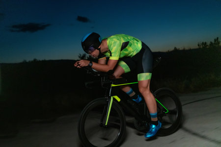 A triathlete rides his bike in the darkness of night, pushing himself to prepare for a marathon. The contrast between the darkness and the light of his bike creates a sense of drama and highlights the athletes determination and perseverance.の写真素材