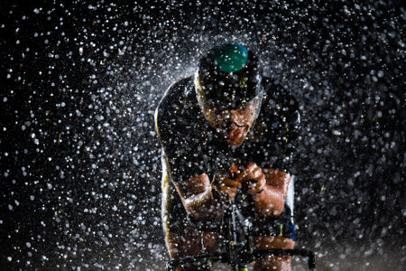 A triathlete braving the rain as he cycles through the night, preparing himself for the upcoming marathon. The blurred raindrops in the foreground and the dark, moody atmosphere in the background add to the sense of determination and grit shown by the athlete.の写真素材