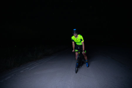 A triathlete rides his bike in the darkness of night, pushing himself to prepare for a marathon. The contrast between the darkness and the light of his bike creates a sense of drama and highlights the athletes determination and perseverance.の写真素材