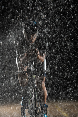 A triathlete braving the rain as he cycles through the night, preparing himself for the upcoming marathon. The blurred raindrops in the foreground and the dark, moody atmosphere in the background add to the sense of determination and grit shown by the athlete.の写真素材