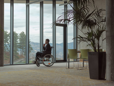 A melancholic businessman in a wheelchair sitting with a sad expression, gazing through the window of a modern office, conveying a sense of solitudeの写真素材