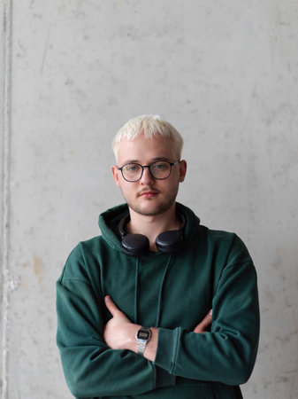 A man with blue hair, eyeglasses, and a green sweatshirt confidently poses with his arms crossed against a gray background, showcasing his fashionable and unique style.の写真素材