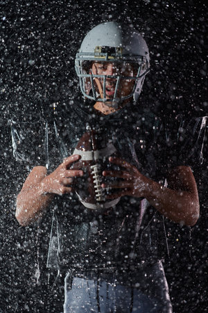 American Football Field: Lonely Athlete Warrior Standing on a Field Holds his Helmet and Ready to Play. Player Preparing to Run, Attack and Score Touchdown. Rainy Night with Dramatic Fog, Blue Lightの写真素材