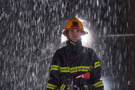 A determined female firefighter in a professional uniform striding through the dangerous, rainy night on a daring rescue mission, showcasing her unwavering bravery and commitment to saving lives.の写真素材