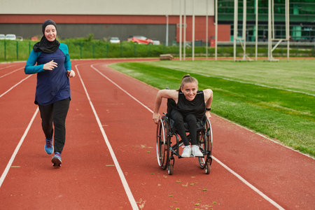 A Muslim woman in a burqa running together with a woman in a wheelchair on the marathon course, preparing for future competitions.の写真素材