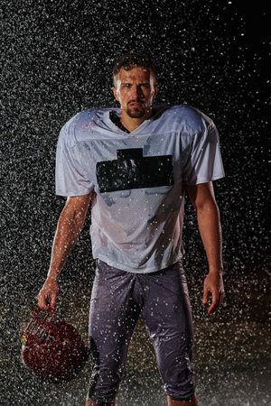 American Football Field: Lonely Athlete Warrior Standing on a Field Holds his Helmet and Ready to Play. Player Preparing to Run, Attack and Score Touchdown. Rainy Night with Dramatic Fog, Blue Lightの写真素材