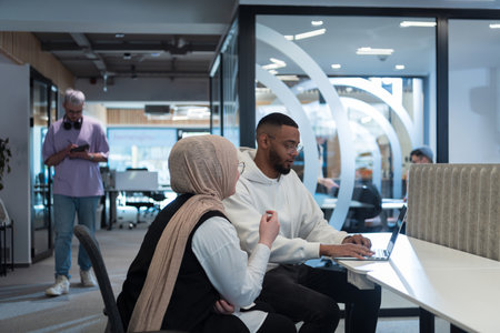 In a modern office setting, an African American businessman and his Muslim colleague, wearing a hijab, engage in collaborative discussions, tackling various business tasks and solving problems together, showcasing diversity, ambition, and teamwork in their dynamic and inclusive workplace.の写真素材