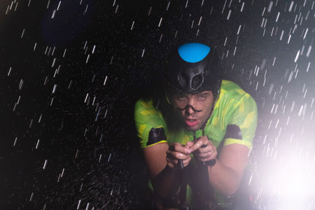 A triathlete braving the rain as he cycles through the night, preparing himself for the upcoming marathon. The blurred raindrops in the foreground and the dark, moody atmosphere in the background add to the sense of determination and grit shown by the athlete.の写真素材