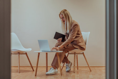 A professional businesswoman sits on a chair, surrounded by a serene beige background, diligently working on her laptop, showcasing dedication and focus in her pursuit of successの写真素材