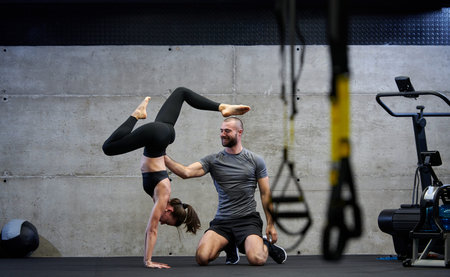 A muscular man assisting a fit woman in a modern gym as they engage in various body exercises and...