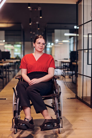 A modern young businesswoman in a wheelchair is surrounded by an inclusive workspace with glass-walled offices, embodying determination and innovation in the business worldの写真素材