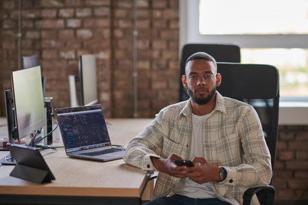 A young African American entrepreneur takes a break in a modern office, using a smartphone to browse...