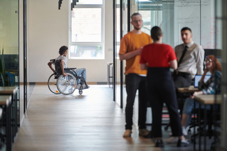 A diverse group of colleagues engages in a discussion about business challenges within a modern coworking startup center, while in the background, their wheelchair-bound colleague symbolizes inclusivity and determination.の写真素材
