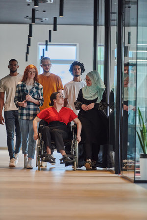 A diverse group of young business people walking a corridor in the glass-enclosed office of a modern startup, including a person in a wheelchair and a woman wearing a hijab, showing a dynamic mix of innovation and unity.の写真素材